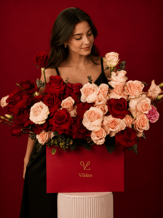 Female model gazes at red and blush roses in a Vilden box on a cream cylinder stand.