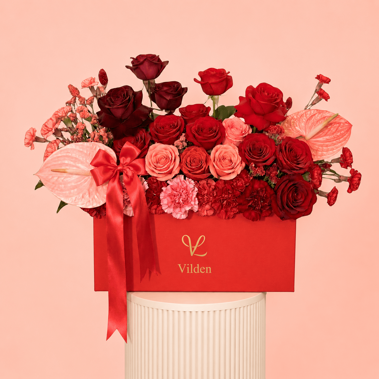 Vilden luxury red and blush flower arrangement in a red box with burgundy roses, red roses, peach roses, pink carnations, pink anthuriums, and a satin red bow, displayed on a cream ribbed pedestal against a soft peach-pink background.