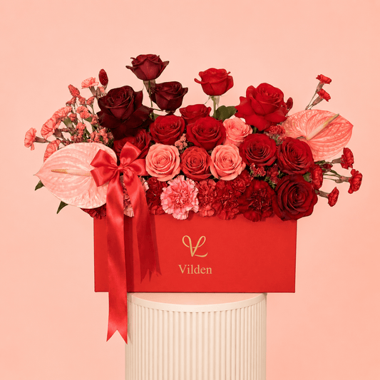 Vilden luxury red and blush flower arrangement in a red box with burgundy roses, red roses, peach roses, pink carnations, pink anthuriums, and a satin red bow, displayed on a cream ribbed pedestal against a soft peach-pink background.
