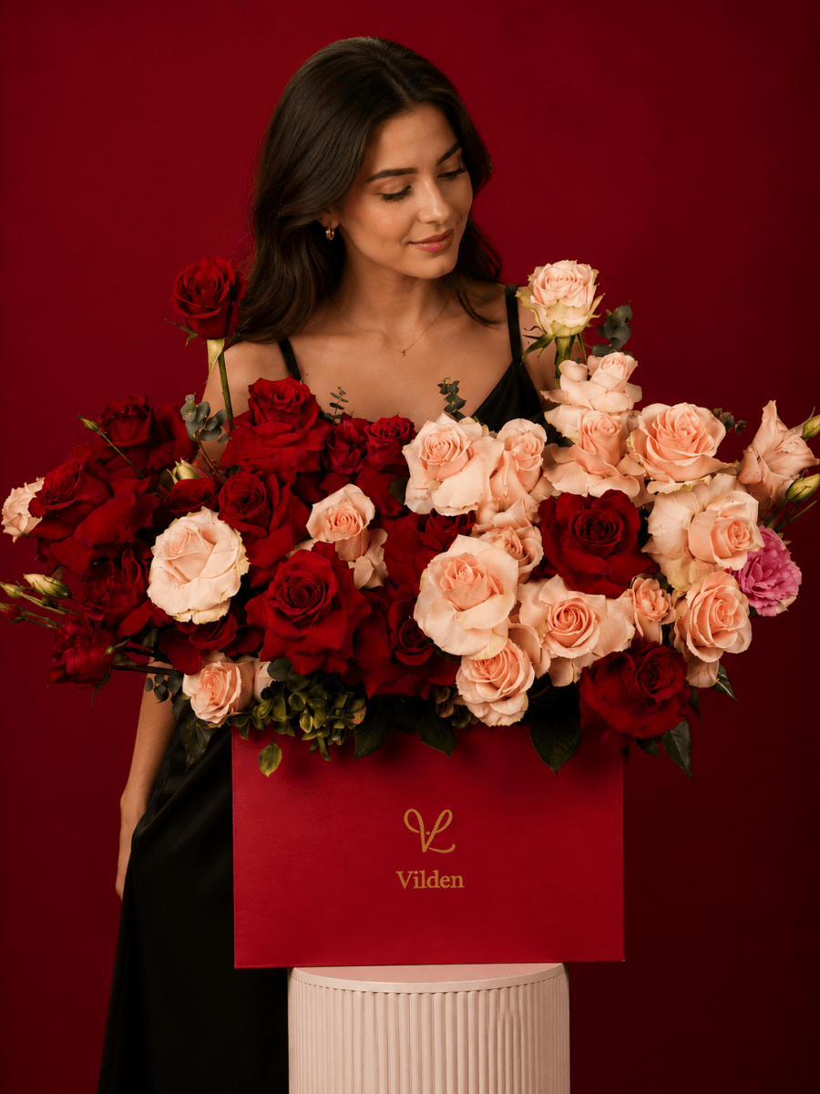 Female model gazes at red and blush roses in a Vilden box on a cream cylinder stand.