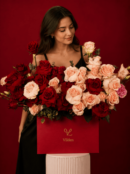 Female model gazes at red and blush roses in a Vilden box on a cream cylinder stand.