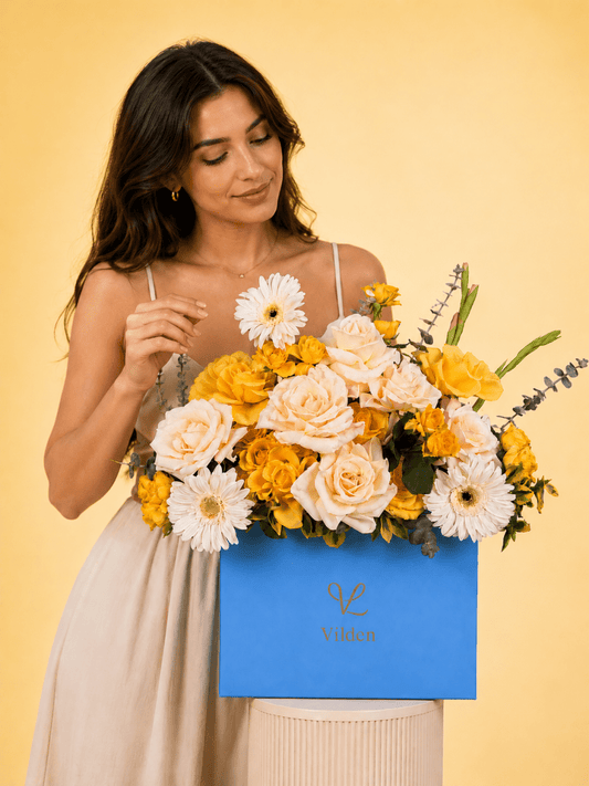 Female model with blue Vilden flower box of yellow roses and white blooms on cream stand.