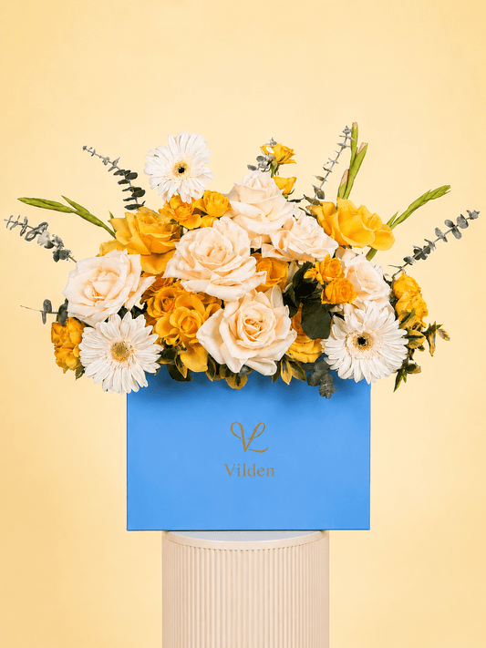 Golden harmony flower arrangement in a blue box with ivory roses, yellow roses, white gerbera daisies, and eucalyptus on a cream ribbed pedestal against a soft light yellow background.