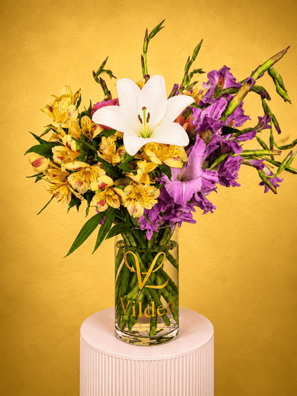 Saffron Mirage Vase Arrangement with yellow alstroemeria, violet gladiolus, ivory lilies, and greenery in a clear Vilden vase