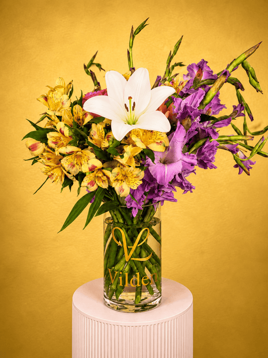Saffron Mirage Vase Arrangement with yellow alstroemeria, violet gladiolus, ivory lilies, and greenery in a clear Vilden vase