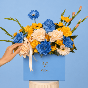 Sun Meets Sky arrangement. Hand gently touches blue, yellow, and cream flowers in a Vilden blue box on a cream stand.