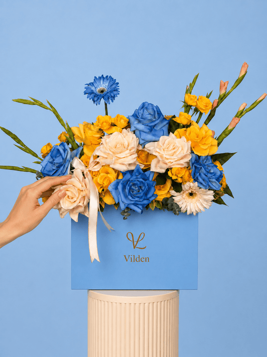 Sun Meets Sky arrangement. Hand gently touches blue, yellow, and cream flowers in a Vilden blue box on a cream stand.