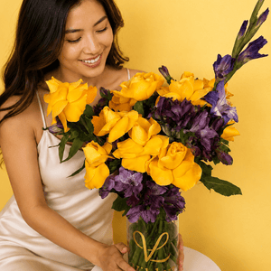 A smiling woman in a cream satin dress holds Sunlit Royale a large golden and violet flower arrangement bouquet