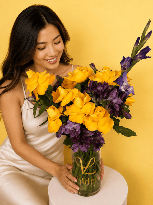 A smiling woman in a cream satin dress holds Sunlit Royale a large golden and violet flower arrangement bouquet