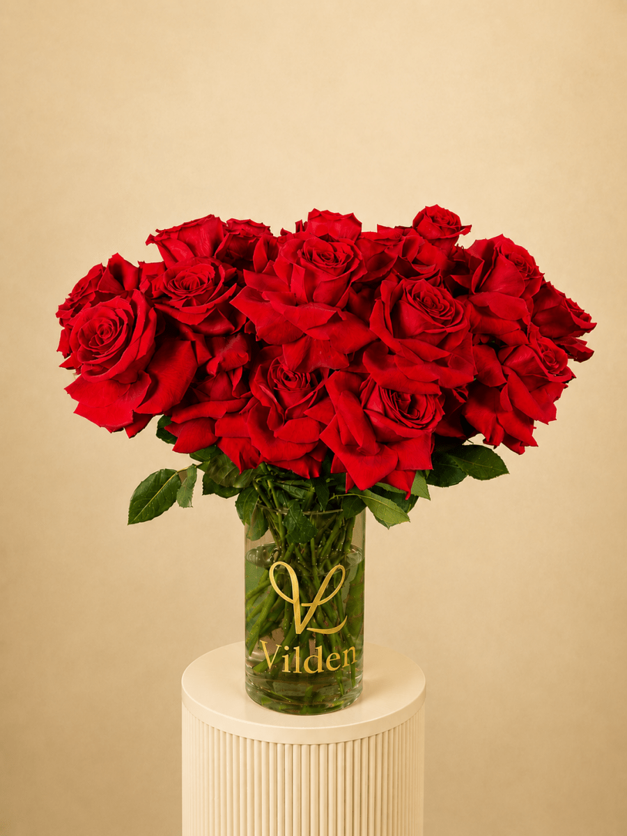 Vilden Rouge arrangement in a clear Vilden vase on a creamy white ribbed pedestal against a soft warm beige background.