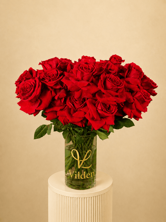 Vilden Rouge arrangement in a clear Vilden vase on a creamy white ribbed pedestal against a soft warm beige background.