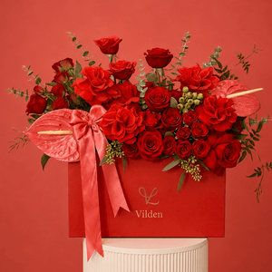 Luxury arrangement in a red box with red roses, coral anthuriums, and a satin pink bow, displayed on a cream ribbed pedestal.