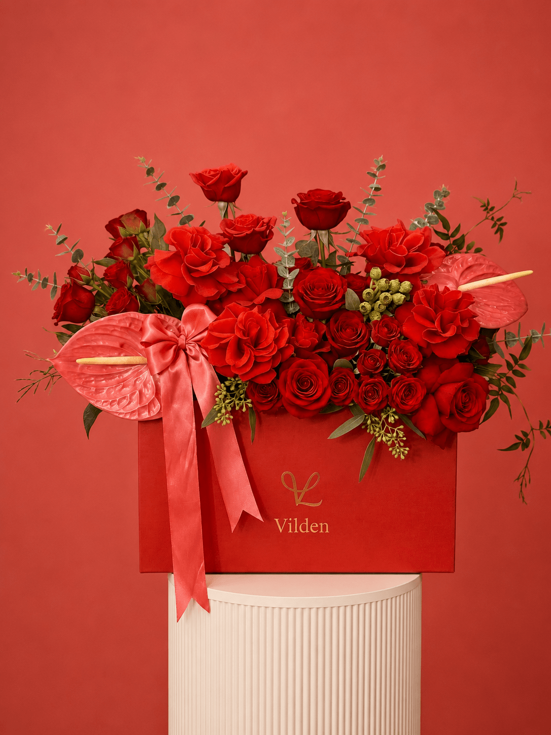 Luxury arrangement in a red box with red roses, coral anthuriums, and a satin pink bow, displayed on a cream ribbed pedestal.