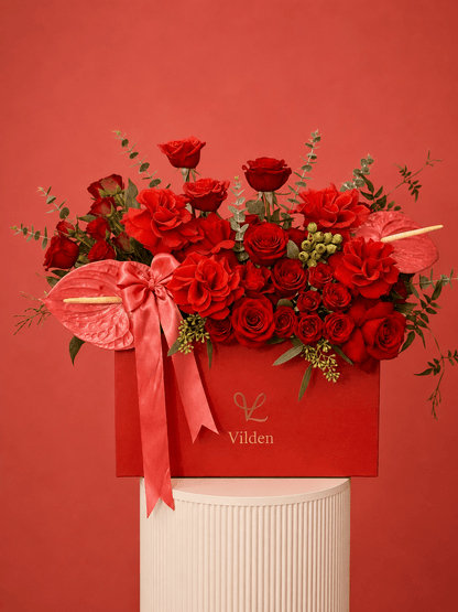 Luxury arrangement in a red box with red roses, coral anthuriums, and a satin pink bow, displayed on a cream ribbed pedestal.