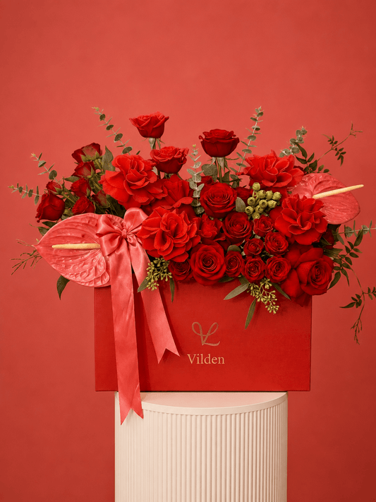 Luxury arrangement in a red box with red roses, coral anthuriums, and a satin pink bow, displayed on a cream ribbed pedestal.