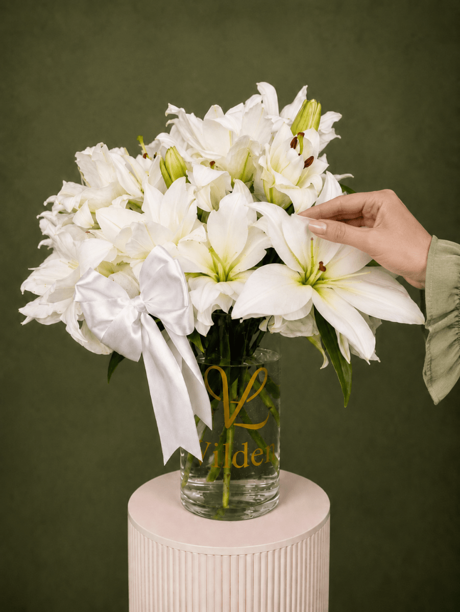 Ivory Lily Reverie with white lilies, unopened lily buds, and a white satin bow in a clear Vilden vase with a soft tough